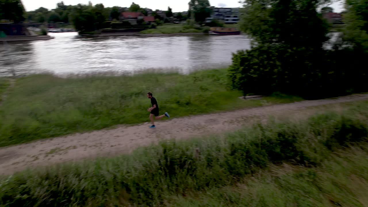 Ripresa aerea che segue un trail runner che parte lungo il fiume IJssel nella pianura olandese, con la sagoma della città turrita di Zutphen sullo sfondo