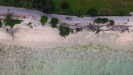 zenithal pan left over a sandy beach, trees, and coastal road with a small vehicle