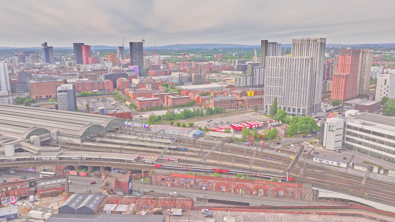 Wide-angle aerial static shot of Manchester city centre showing the train station, modern high-rises, and industrial heritage structures with cloudy sky and urban infrastructure in the foreground