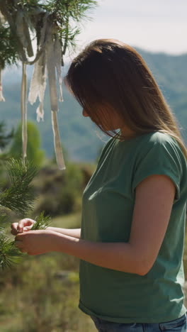young woman ties bow to make wishes on spruce tree twig at countryside side view. traditional ritual for luck. lady near wishing tree at highland