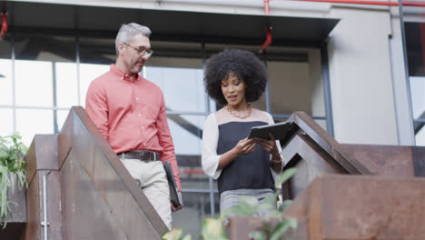 Diverse-male-and-female-colleague-walking-down-stairs-with-tablet-and-laptop,-talking,-slow-motion