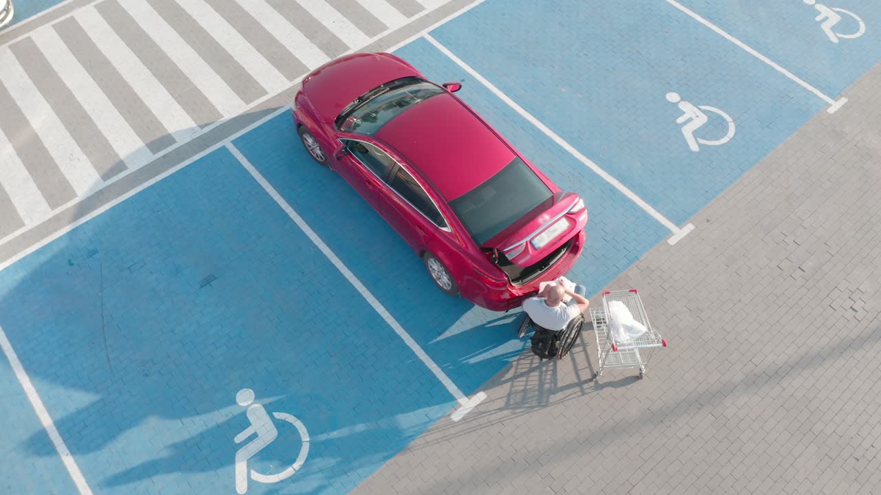 체어에 앉은 장애인 성인 남자가 슈퍼마켓 주차장에서 차의 트크에 물건을 넣고 있다.