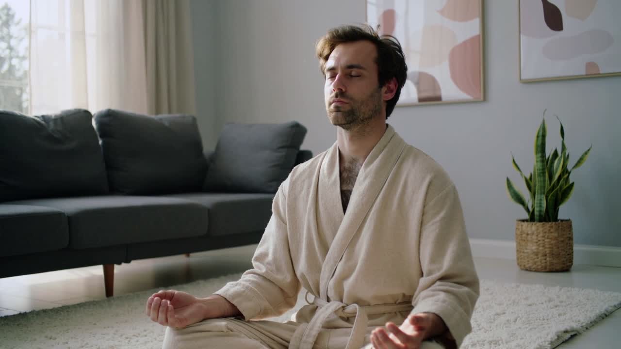 Uomo seduto in accappatoio beige che medita in mudra a casa, con luce soffusa che entra dalla finestra, cercando la calma.