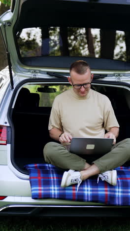 caucasian man sitting in the trunk of his car puts on glasses. freelancer is getting ready to work on his laptop. beautiful summer nature at backdrop. vertical video