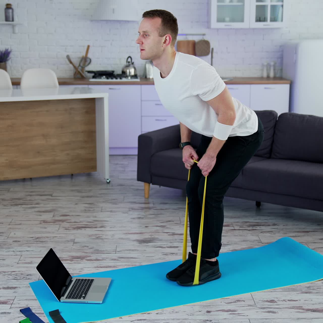 Concentrated man exercising with a rubber band at home. Young man doing working out while standing on a mat in the kitchen. Sportive lifestyle during quarantine