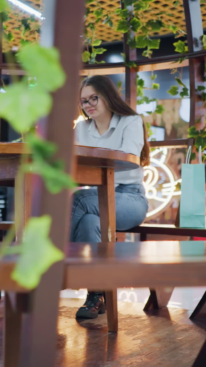 장식 좌석에 앉아있는 젊은 여성, 그녀의 에 분홍색과 민트 녹색 쇼핑 가방과 함께 그녀의 전화에 초점을 맞추고, 배경에 반이는 불빛, 누군가가 활기찬 분위기에서 뒤를 지나가는 동안