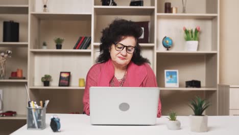 anciana, con una expresión triste, termina su trabajo en el portátil y, quitándose las gafas, se masajea el cuello