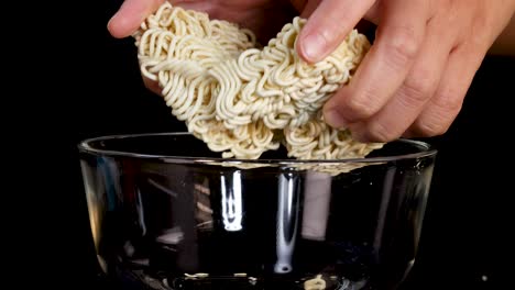 hands break a dry instant noodle block and place it into a clear glass bowl against a black background, with bright, even studio lighting and steady camera