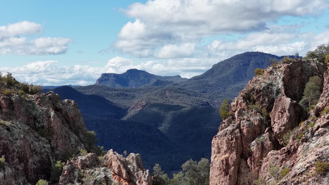 Camera pans across rugged rocky outcrops and forested valleys under bright daylight, revealing expansive mountain scenery in Warrumbungle National Park, Australia