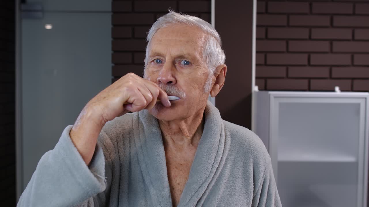 Senior man grandfather in bathrobe brushing teeth, looking into mirror. Morning hygiene at bathroom