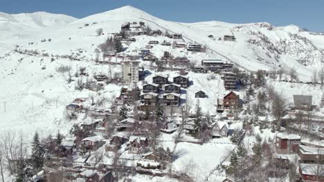 Aerial-orbit-establishing-the-mountain-village-of-Farellones-full-of-snow-in-the-winter-season,-parallax-effect-with-the-Andes-mountain-range-and-El-Colorado-ski-resort,-Chile