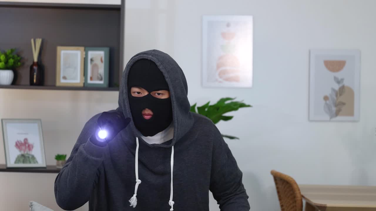 Ein maskierter Eindringling mit grauem Hoodie durchsucht ein Wohnzimmer mit einer hellen Taschenlampe