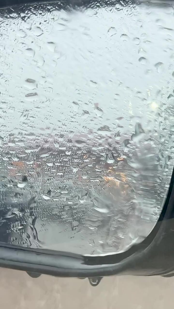 Primo piano di goccioline d'acqua e condensa su uno specchietto retrovisore di un veicolo durante una giornata piovosa. Texture cinematografiche per narrazioni di viaggio e legate al meteo.