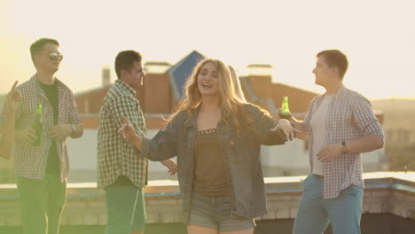 the beautiful american girl is dancing on the roof with her five friends on the party who drinks beer from green bottles. she smiles and enjoys the time in shorts and a light denim jacket in summer evening. her blond hair is flying in the wind.