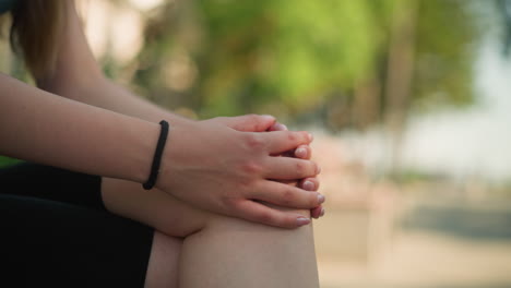 Close-up of young woman seated outdoors with leg crossed, hands clasped together resting on knee, wearing black hand bangle, under soft natural light with blurred green background