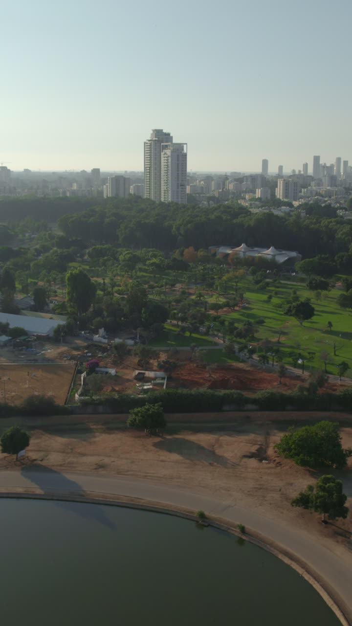 An aerial view in a calm afternoon over the hippopotamus lake of the Ramat Gan Safari when it is empty of visitors. Tel Aviv city towers In the background - sliding shot - vertical video