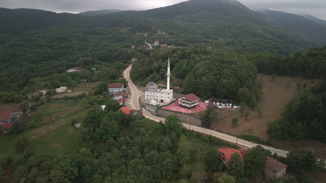 Bulutlu bir günde yeşil tepelerle çevrili Mecidiye köyündeki geleneksel bir Türk camisinin havadan dairesel çekimi.
