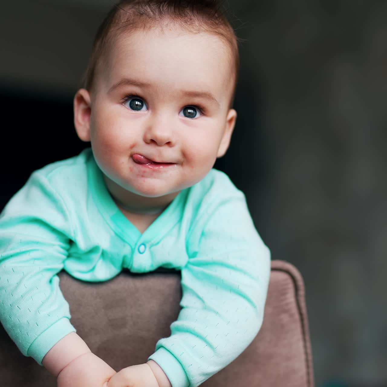 Adorable playful baby boy standing on a chair leaning on chair back. Funny toddler showing tongue smiling