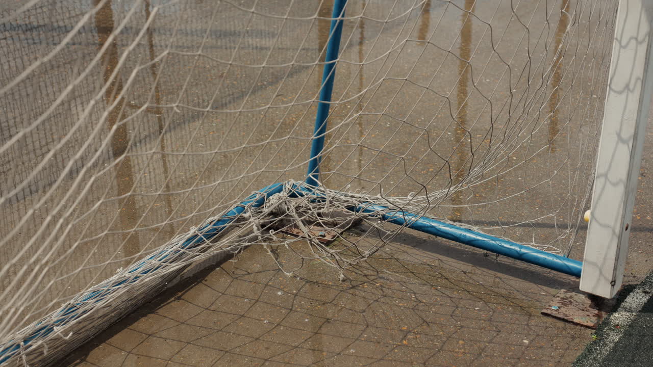 Primo piano di un angolo bagnato di una porta da calcio sotto la pioggia, rete e palo di metallo con vernice blu scrostata, gocce di pioggia che schizzano in una pozzanghera fangosa sul campo, cielo nuvoloso che proietta una luce grigia, stadio vuoto
