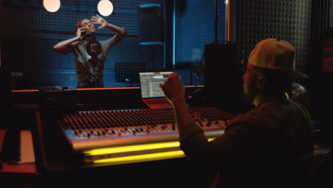 young musical producer sitting in the dark sound studio at the mixer while young female singer singing and recording a song over the glass