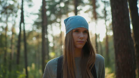 young woman wearing blue headscarf walks through forest, adjusting her backpack and looking contemplatively into distance, sunlight filters through trees, casting a warm glow