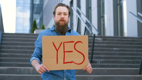 portrait d'un homme caucasien barbu tenant une pancarte "oui" et regardant la caméra dans la rue"