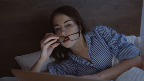 mujer joven cansada y soñolienta que trabaja tarde en la noche en la computadora portátil en la cama, luego se quita las gafas y bosteza