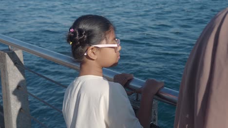 a young girl with pigtails and glasses looks out at the ocean from a pier