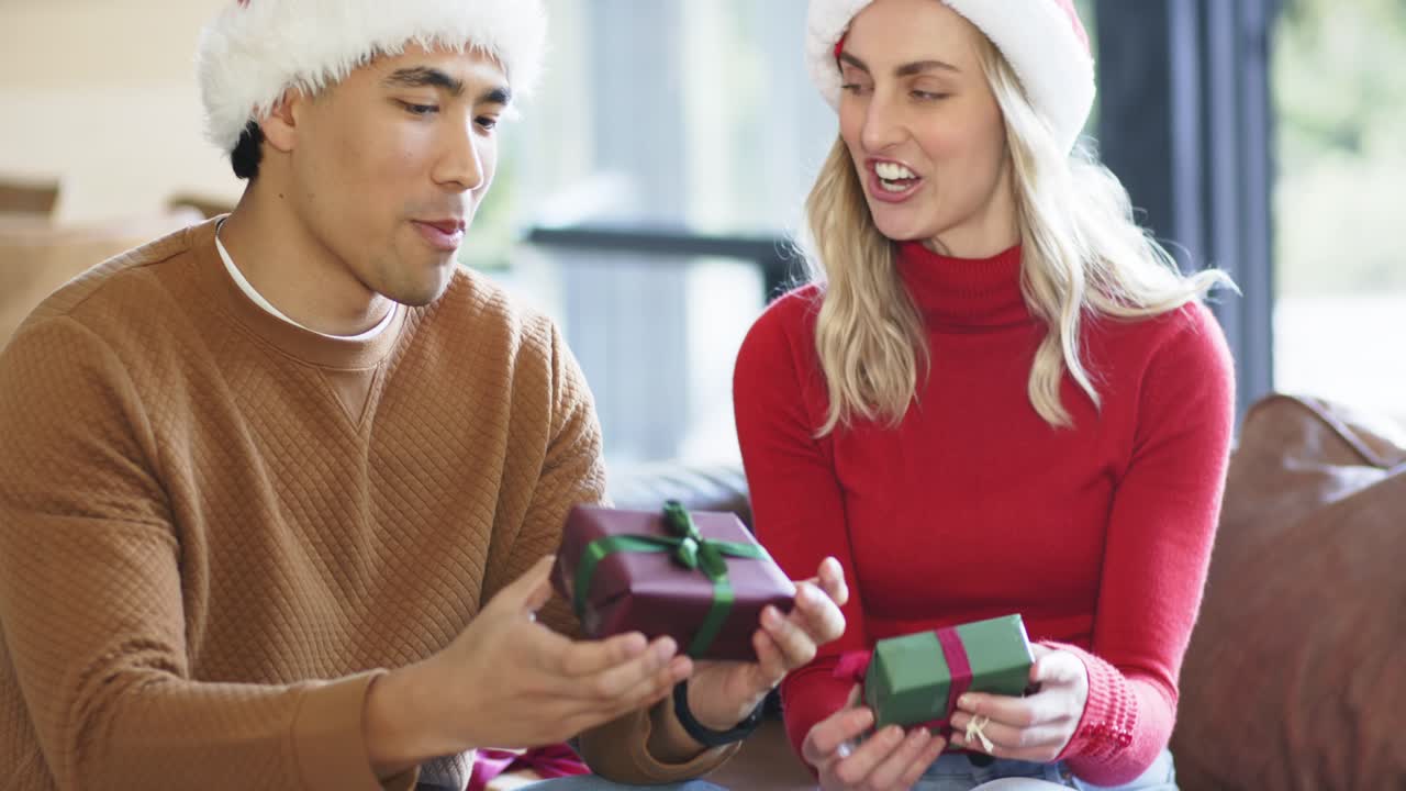 Coppia con regali e cappelli da Babbo Natale che mostra un nastro, festeggiando lo scambio di regali sul divano