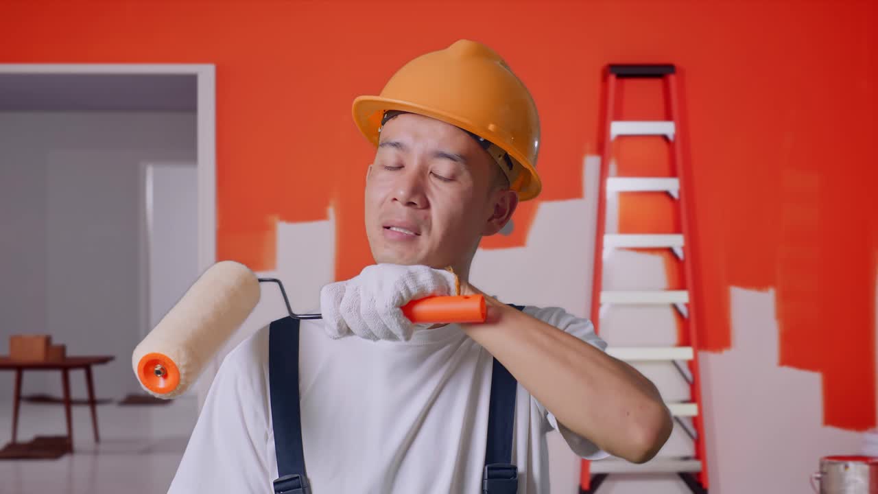 close up de un pintor asiático que usa un casco de seguridad limpiando el sudor y estando cansado mientras está de pie con la pared de pintura en la habitación