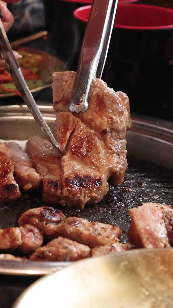 Des mains utilisent des pinces métalliques et des ciseaux pour trancher du porc grillé juteux sur un gril chaud.