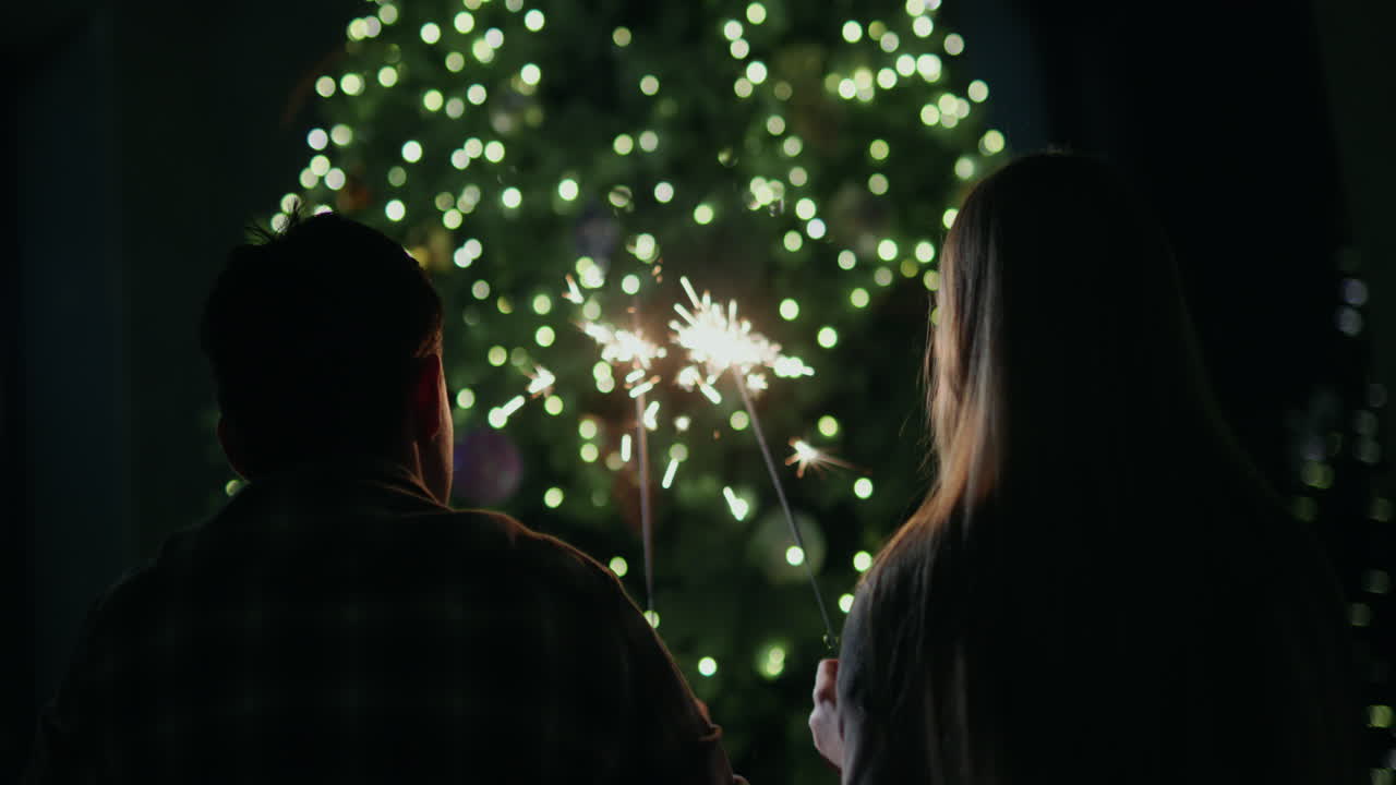 una pareja joven sostiene chispas ardientes en sus manos, se sienta frente a un gran árbol de navidad en casa