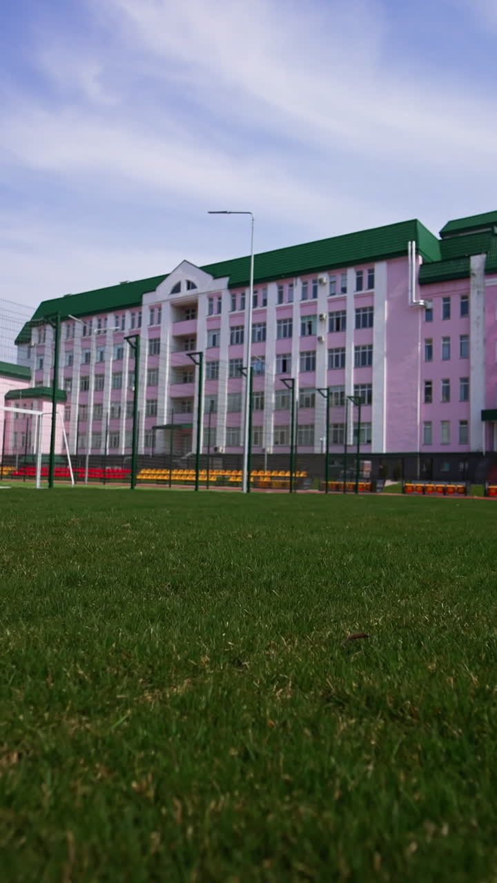 Red running track, green grass and football field located in front of the beautiful pink building. Low angle view. Blue sky at backdrop. Vertical video