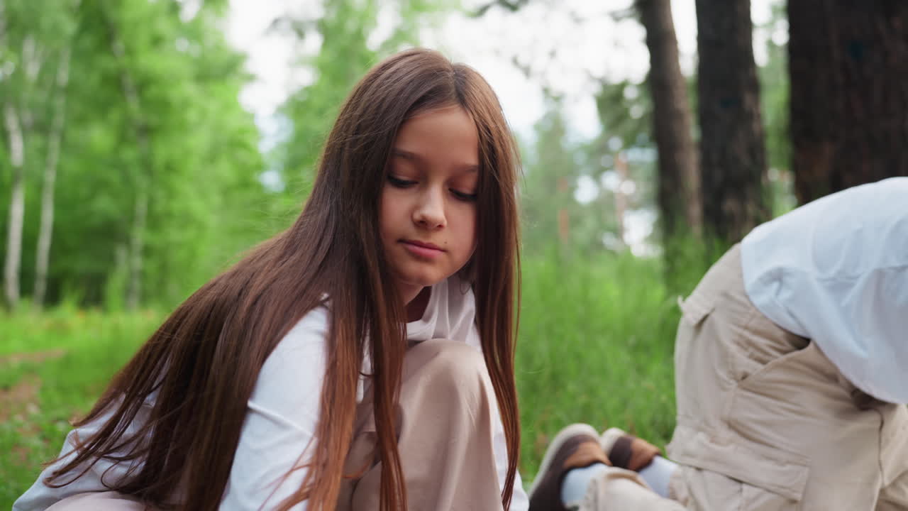 흰 셔츠를 입은 어린 소녀가 숲 바닥에 보라색 천을 펼치고, 주변은 푸르른 나무와 햇빛으로 둘러싸여 자연스러운 움직임, 초점, 부드러운 야외 분위기를 보여주는 미디엄 샷
