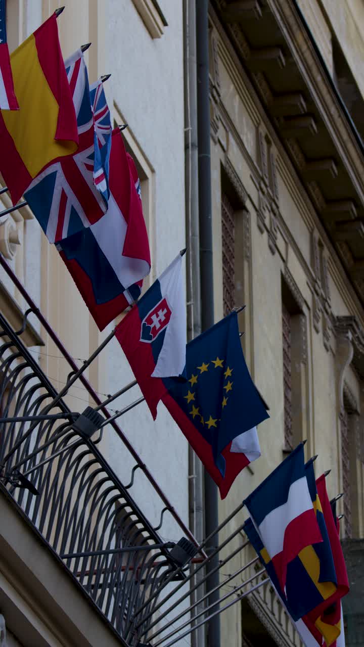 Multiple national flags wave outside ornate embassy facade in Prague’s old town, daylight, steady shot