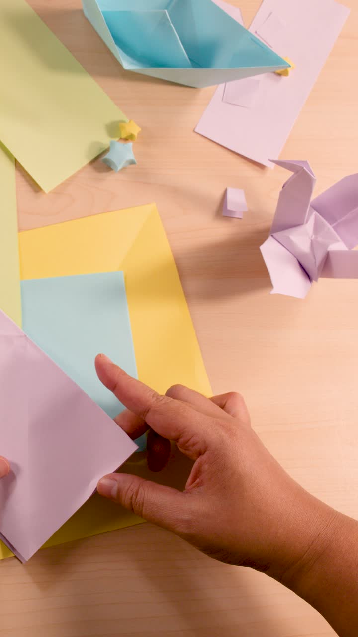 Una persona piega carta pastello per origami su un tavolo di legno, illuminazione soffusa, inquadratura dall'alto.