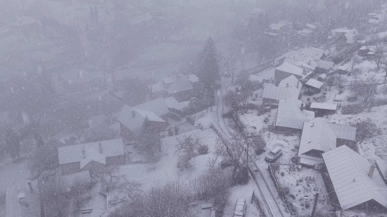 Drone view of a quiet mountain village covered in fresh snow during a heavy winter storm. Houses, gardens, and narrow roads fade into a cold, misty atmosphere