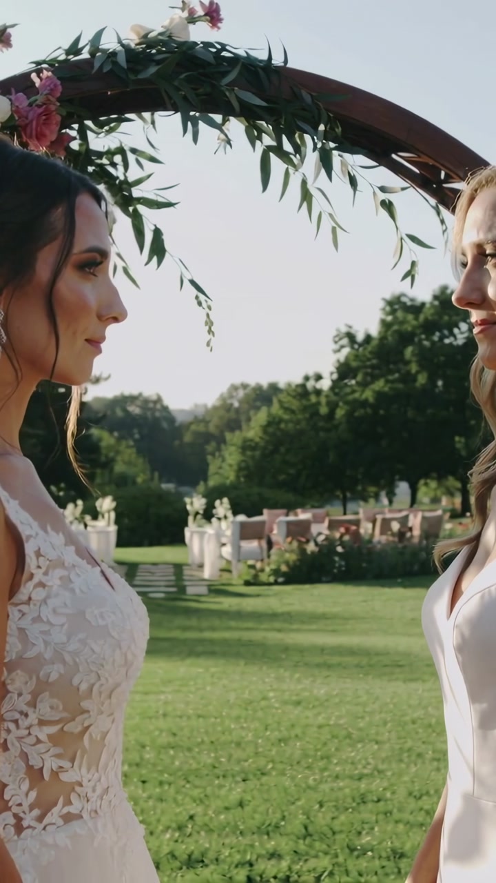 Vertical video: Camera pulling back brides in gowns holding gaze before vows under floral arch