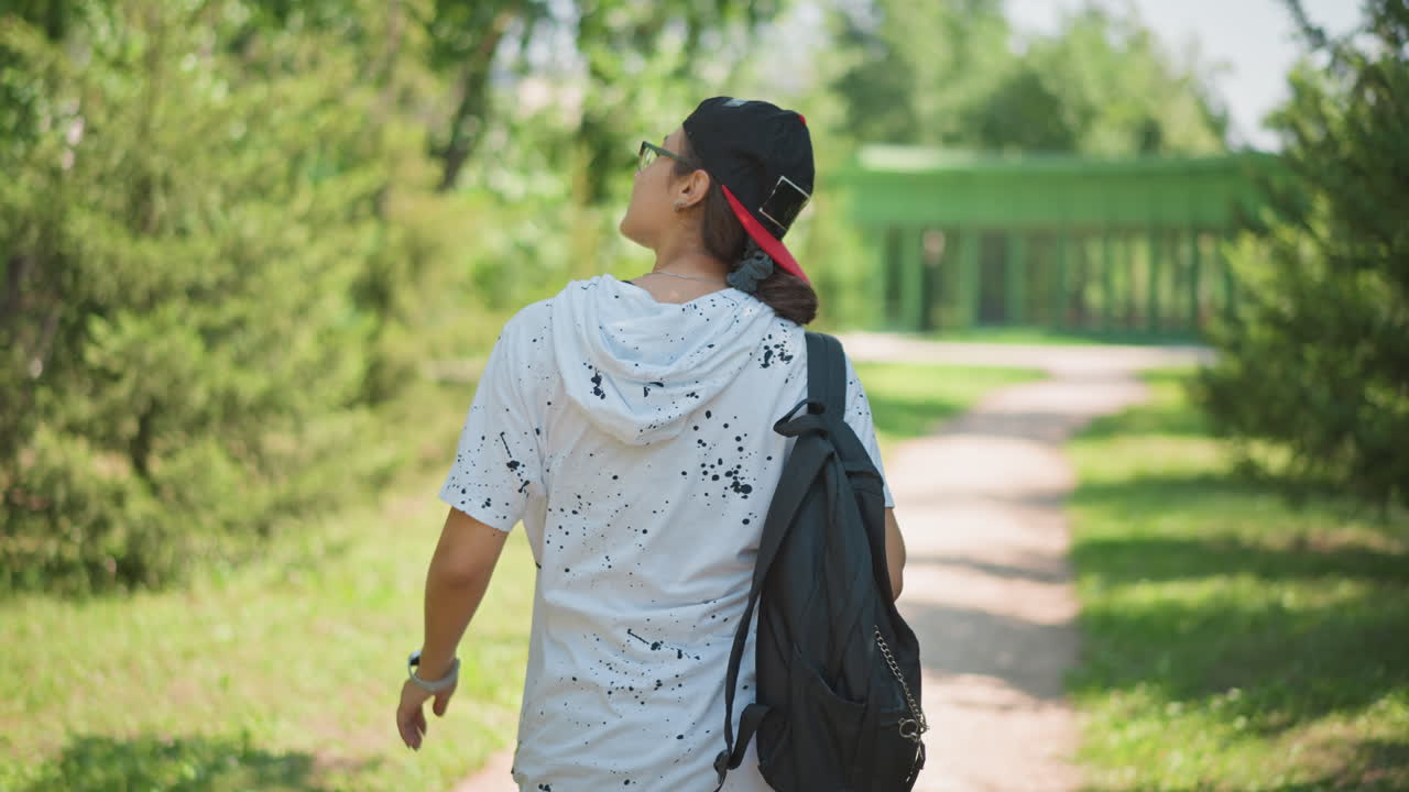 young person walking away trail backpack red cap rear view adjusting strap scanning surroundings summer afternoon leafy corridor serene solitude motion away from camera campus exit casual hoodie