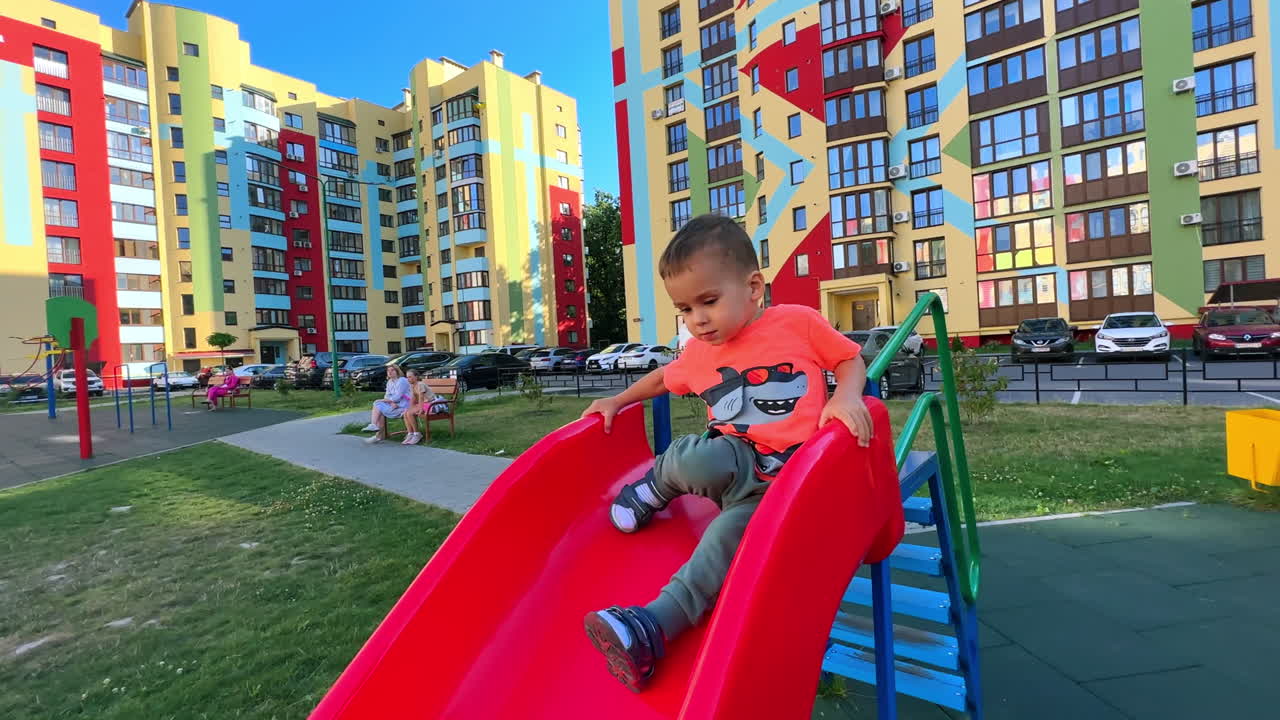 Caucasian toddler climbs on the slide. Baby boy slides down quickly. Bright residential houses at backdrop.
