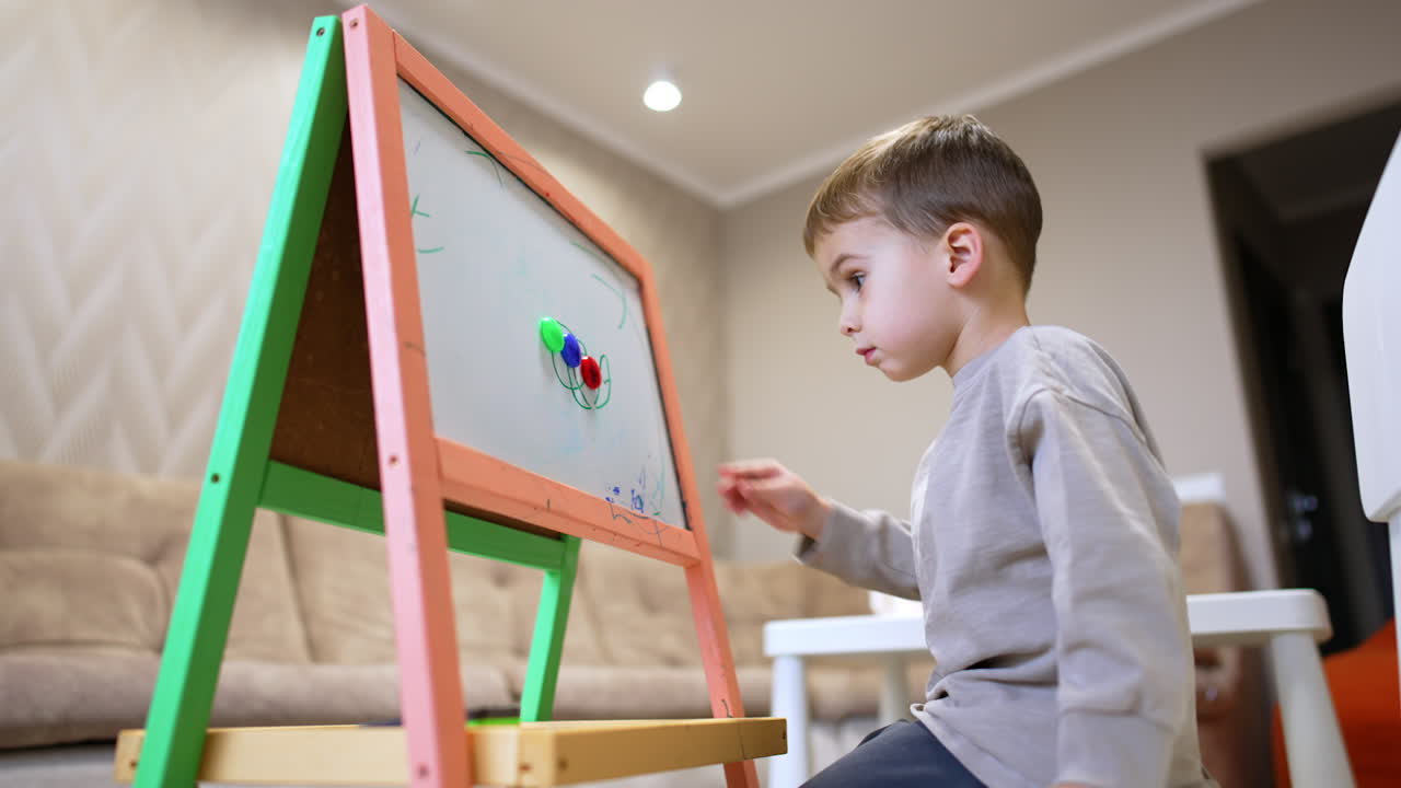 Adorable Caucasian baby boy sits in front of a whiteboard. Low angle view at the kid drawing.