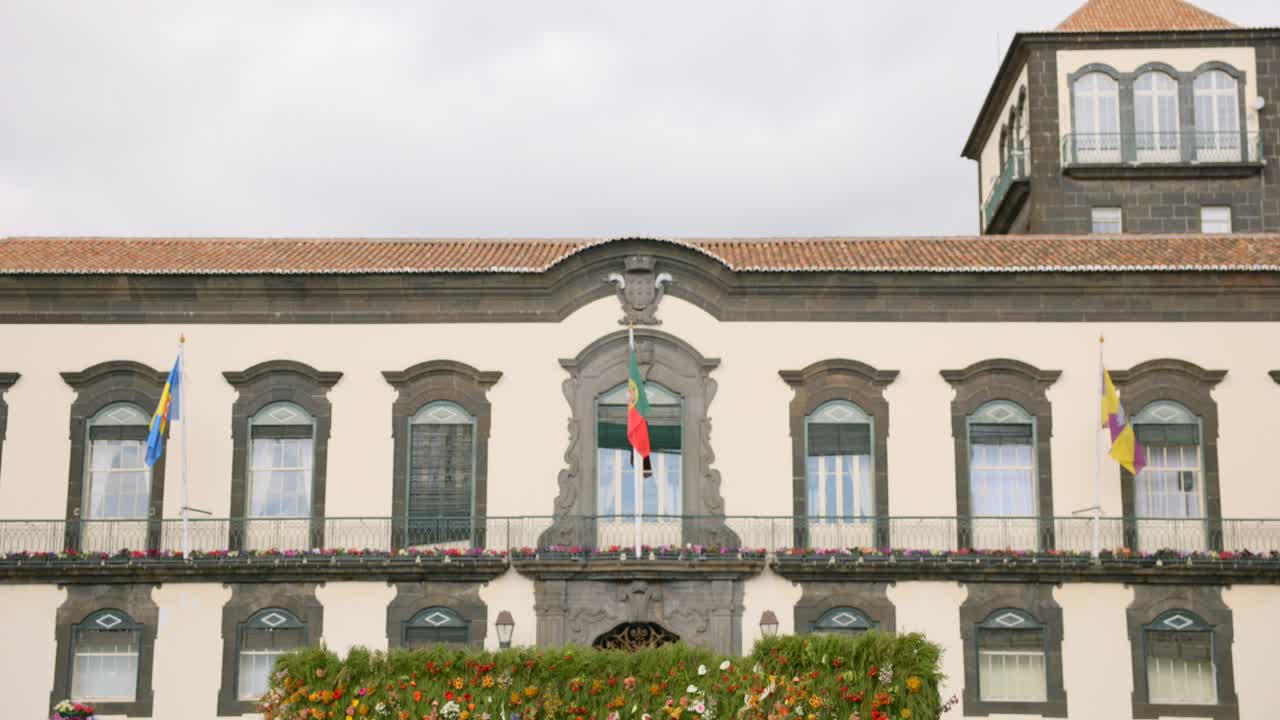 Tilt-up shot revealing a vibrant flower wall installation in front of the Funchal City Hall during the Madeira Flower Festival, with historical architecture and flags in the background
