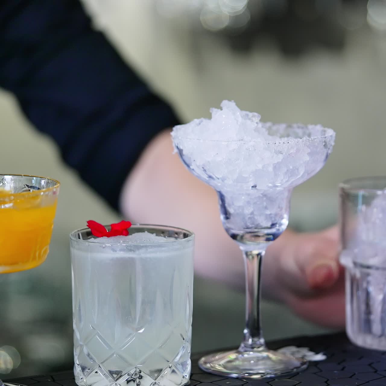 Beautiful diverse glasses filled with ice and drinks. Barman in black shirt preparing cocktails at backdrop in blur