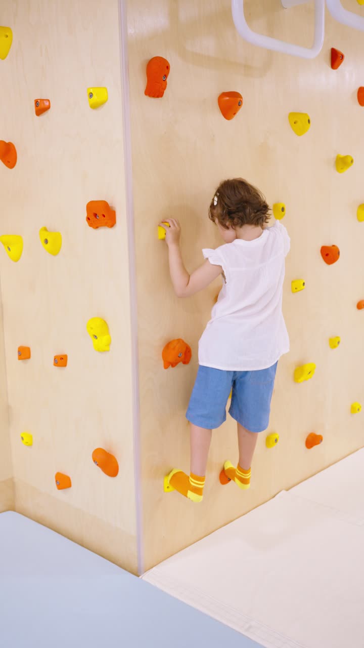 A determined little girl practices her balance and strength by climbing on a colorful bouldering wall at a modern indoor playground, developing her motor skills