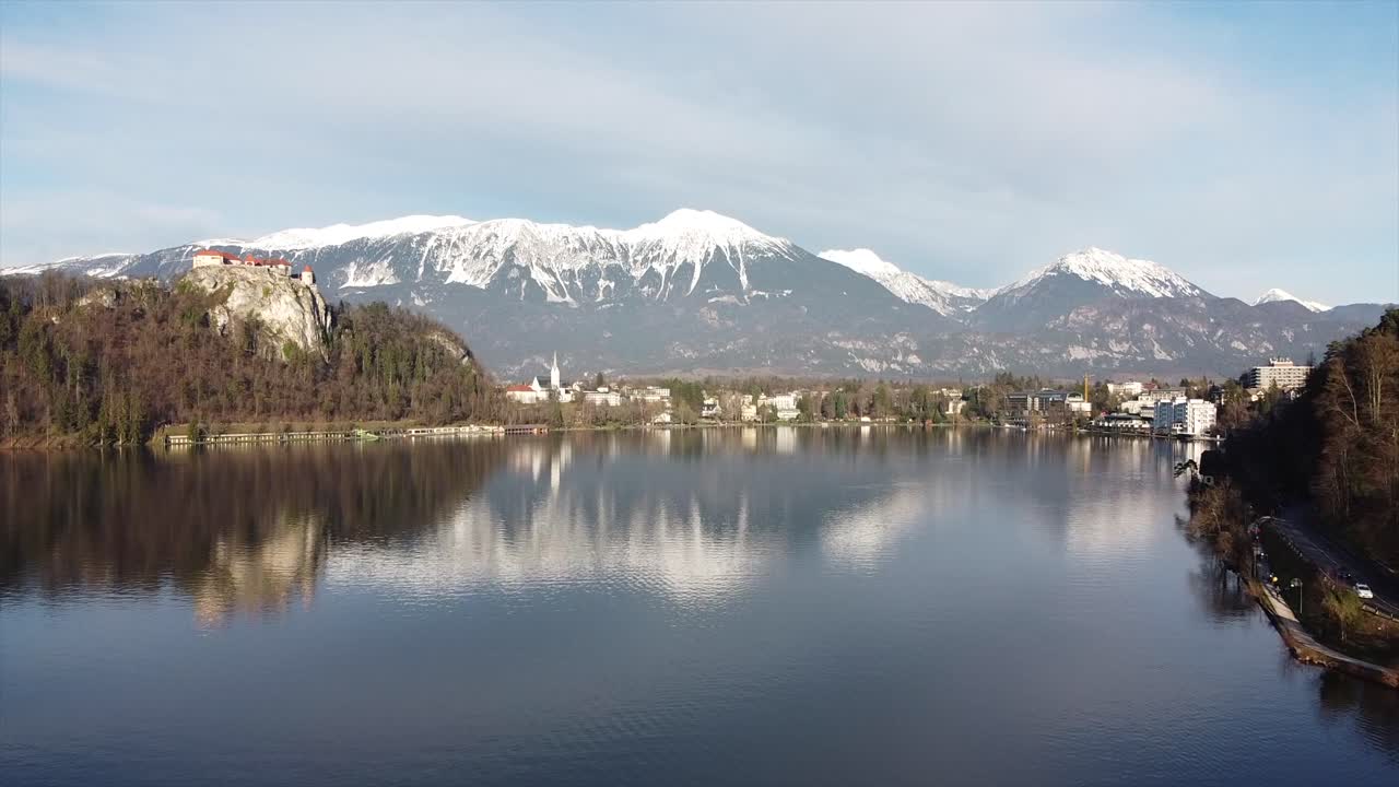 Drone view captures calm lake with reflections of snow-covered mountains and nearby buildings in Canada, showing peaceful water, natural beauty, distant peaks, and clear sky in winter light.