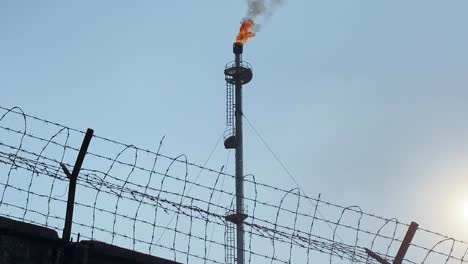 vista en ángulo bajo de la refinería de pilas de bengalas detrás de una cerca cableada, la inclinación hacia arriba revela una llama ardiente y una pila de humo