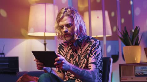 young stylish caucasian man with long hair in glasses sitting on a chair and using a tablet in a retro party at home