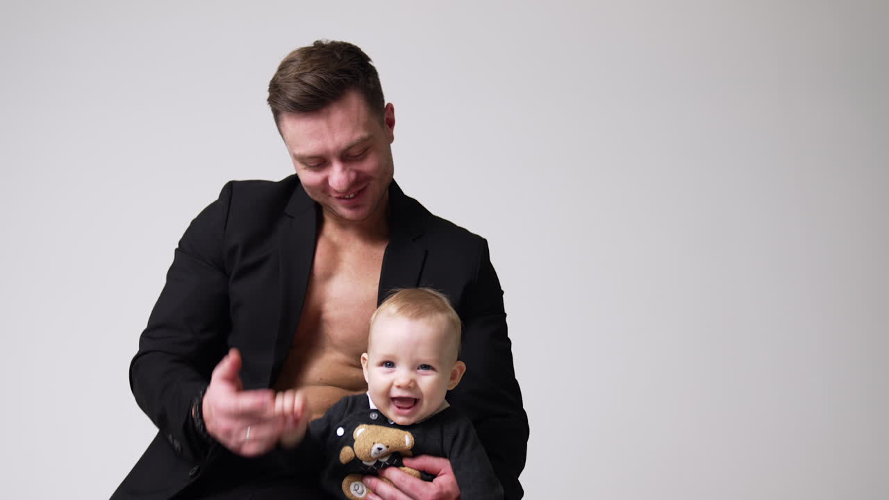 Happy Caucasian man in black jacket holding a cute smiling baby. Father and son smiling. Studio footage. White backdrop.