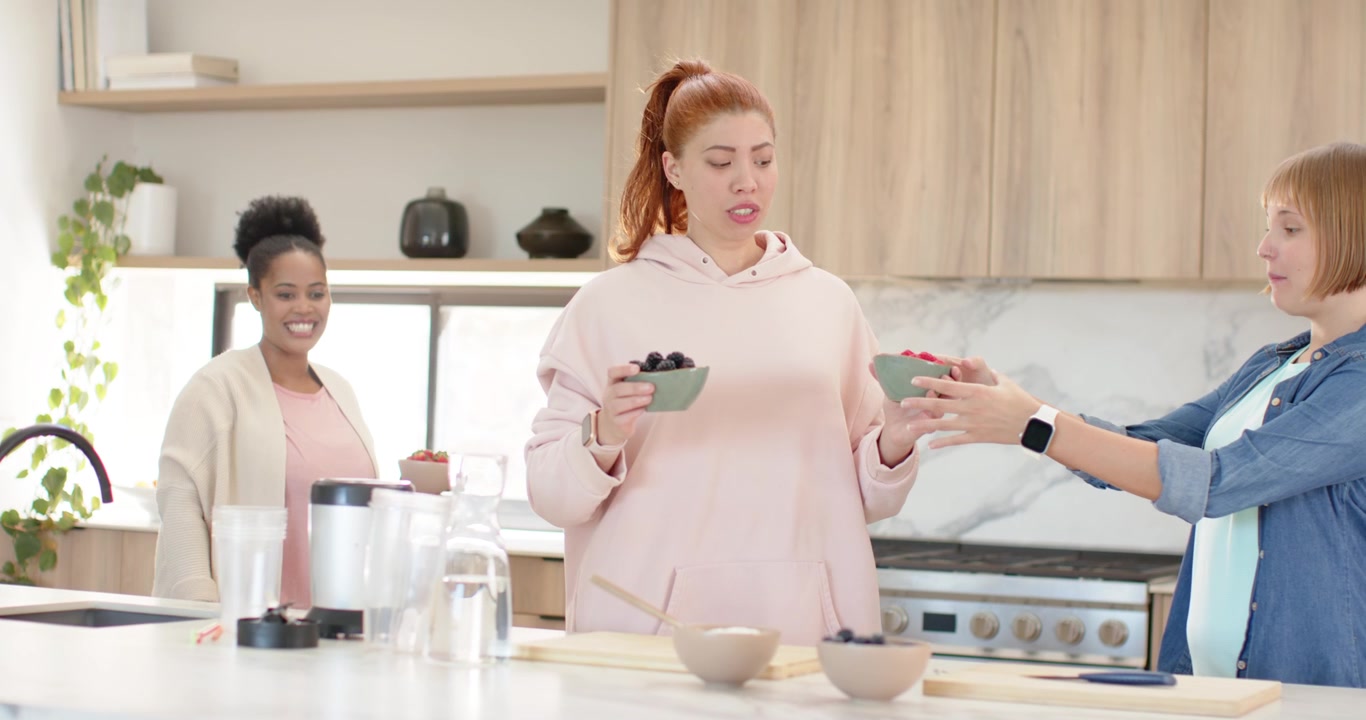Donne diverse che mettono frutta in una cucina di casa mentre una rossa entra tenendo ciotole verdi per servire.