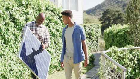 padre afroamericano sosteniendo panel solar hablando con su hijo adulto en un jardín soleado, cámara lenta